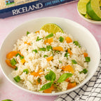 White bowl of rice with vegetables on a pink surface, Riceland rice package in the background.