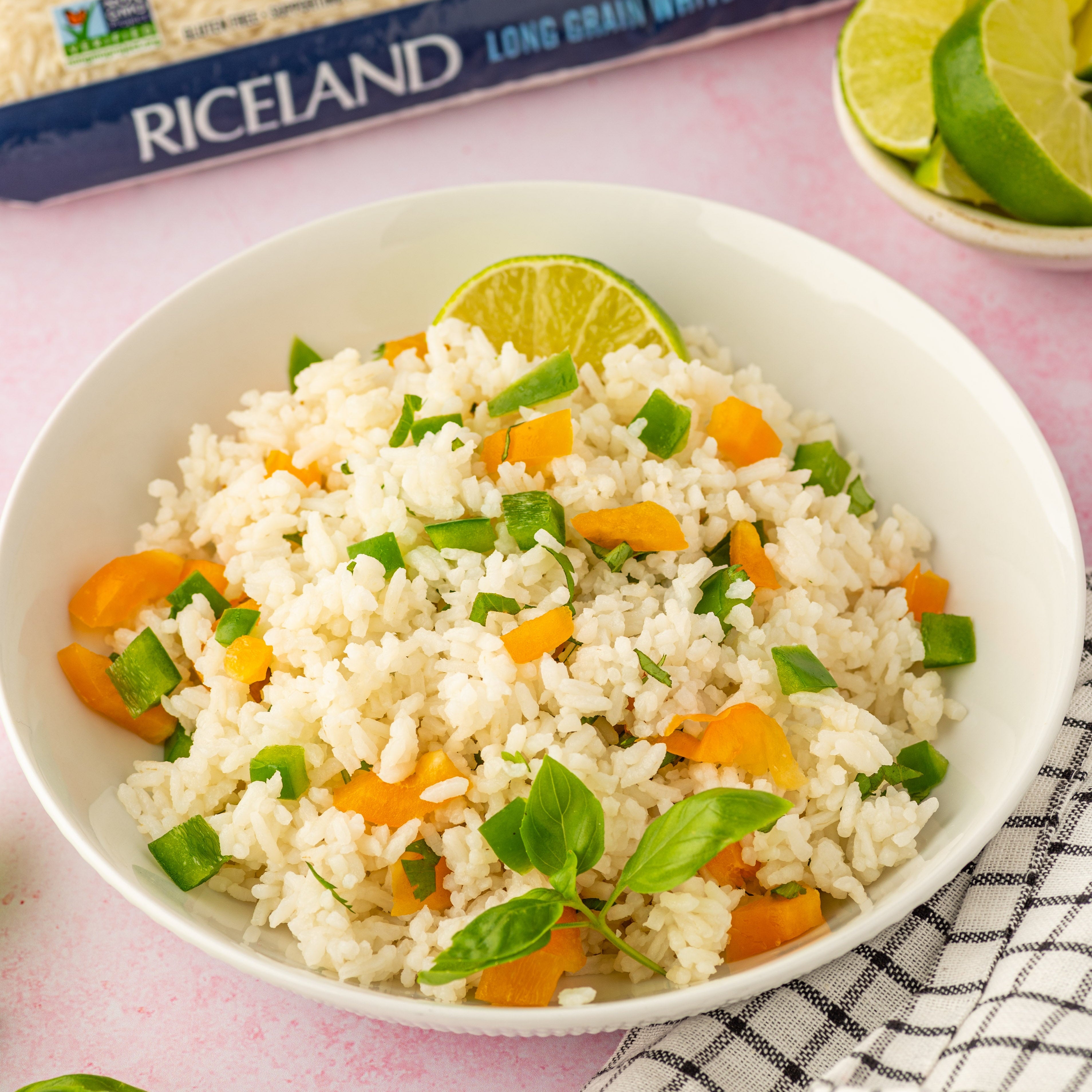 White bowl of rice with vegetables on a pink surface, Riceland rice package in the background.